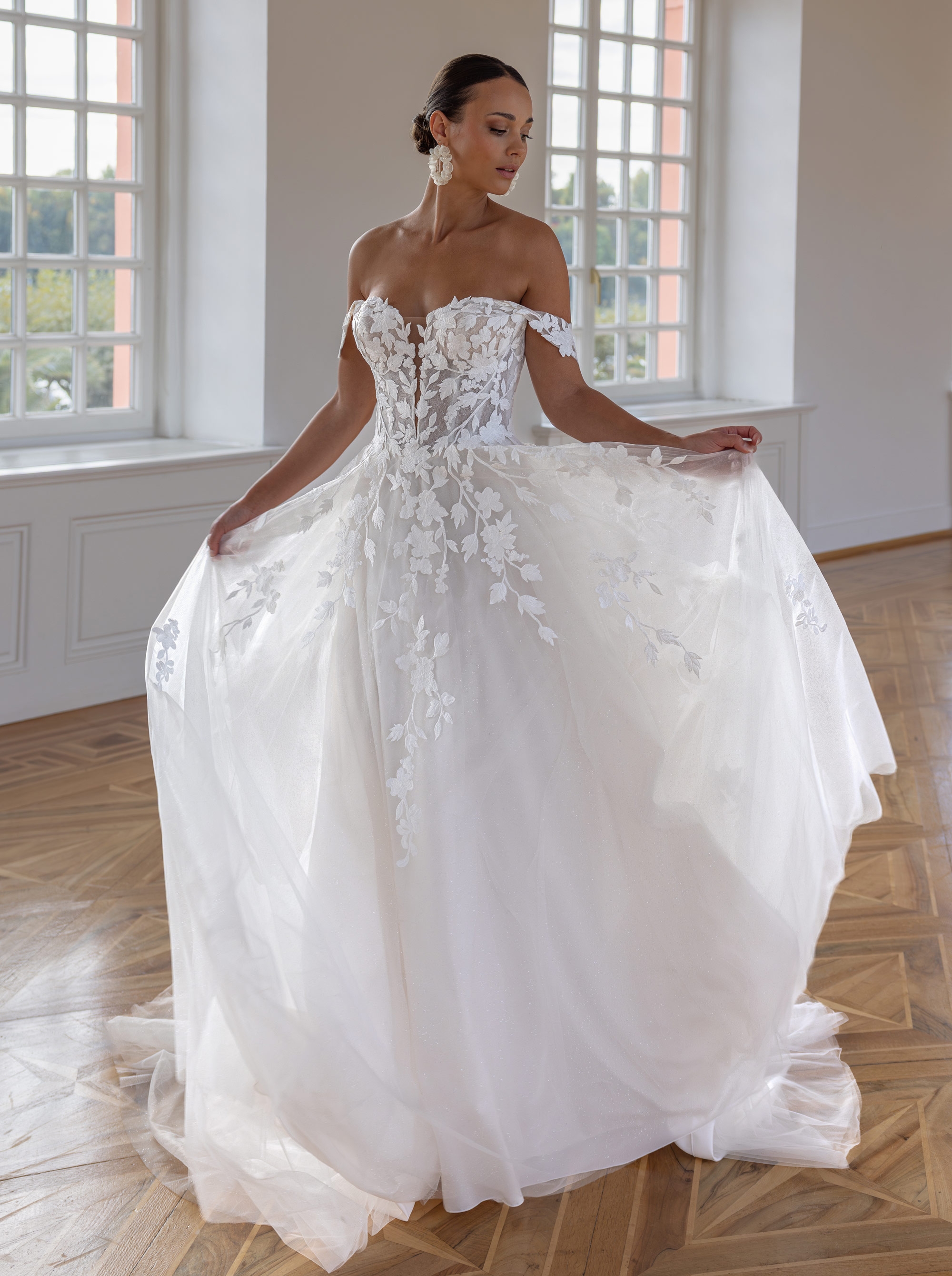 Frau in elegantem, schulterfreiem Brautkleid mit Spitzenblumen im hellen Raum mit Fenster.