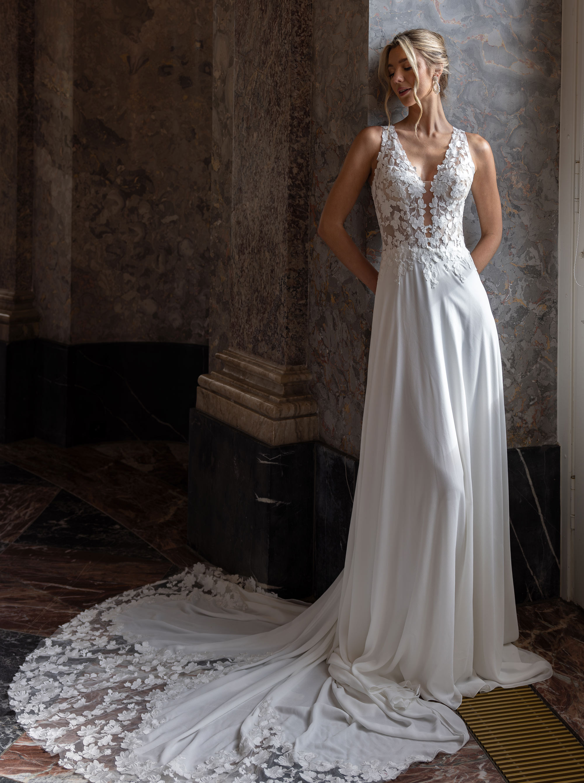Frau in elegantem weißen Hochzeitskleid mit Spitzenoberteil lehnt an einer marmornen Wand.