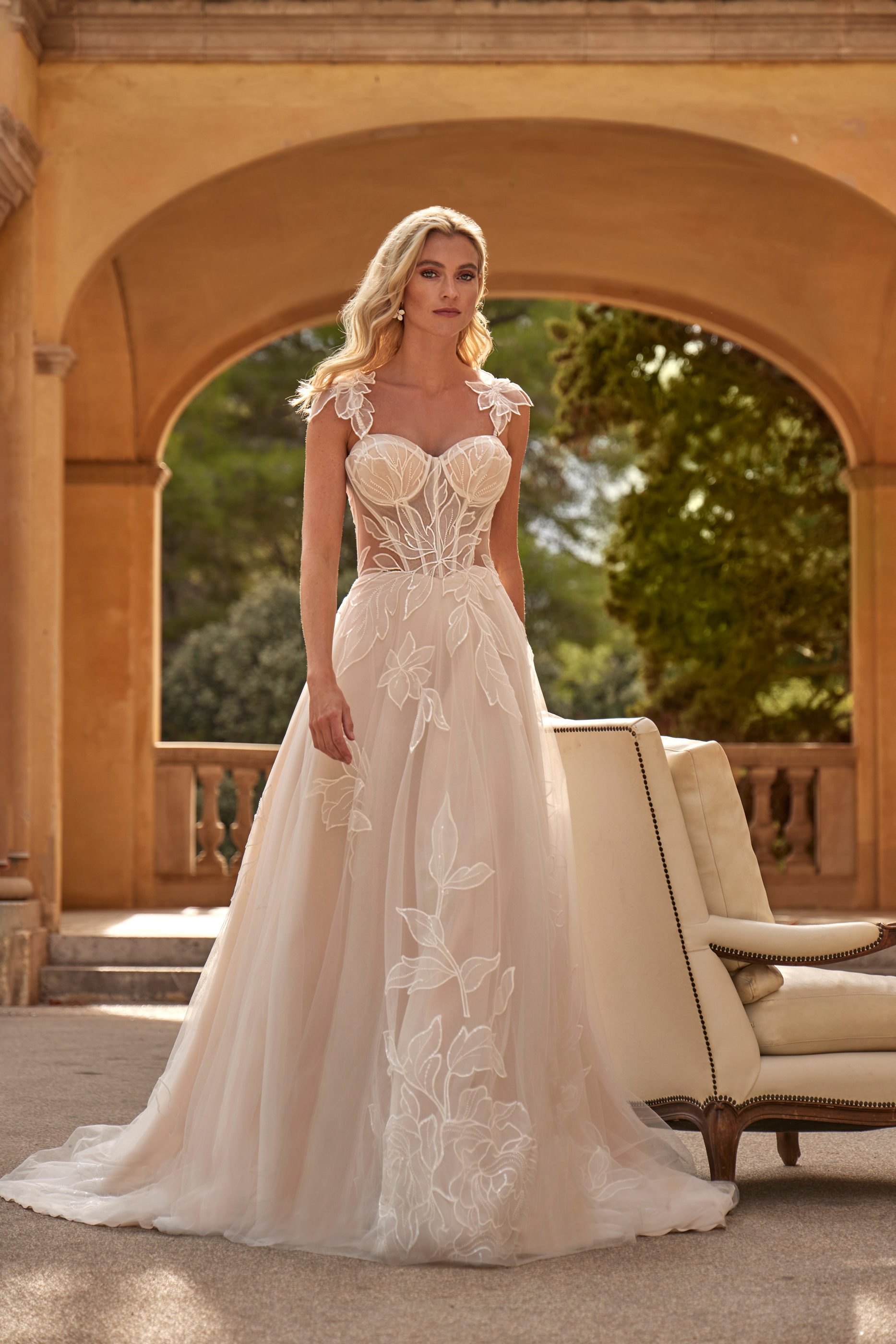 Frau im weißen Brautkleid mit floralen Stickereien steht vor einem Arkadenbogen und einem Stuhl.