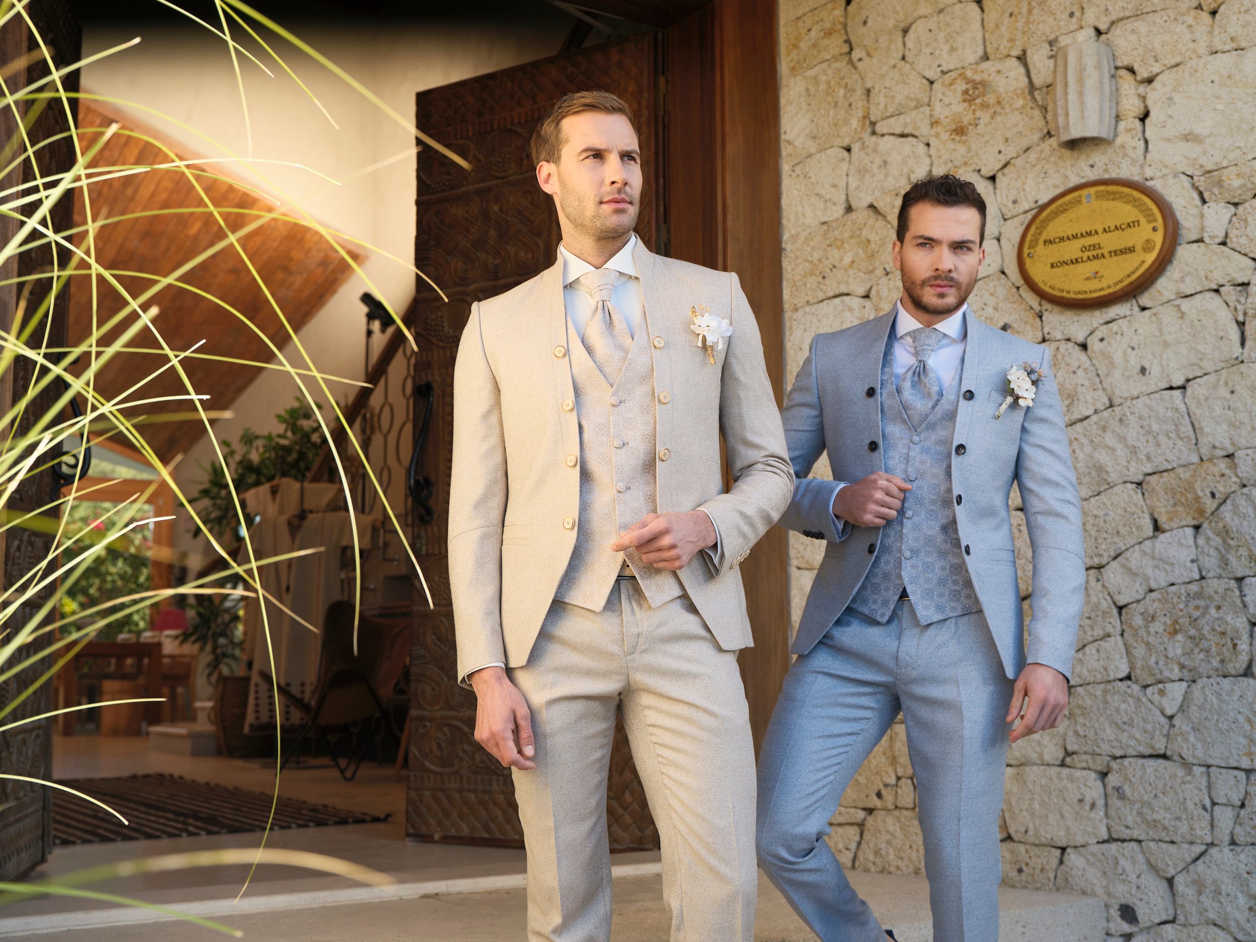 Zwei Männer tragen elegante Hochzeitsanzüge, einer beige, der andere hellblau, vor einem Steinmauer-Eingang.