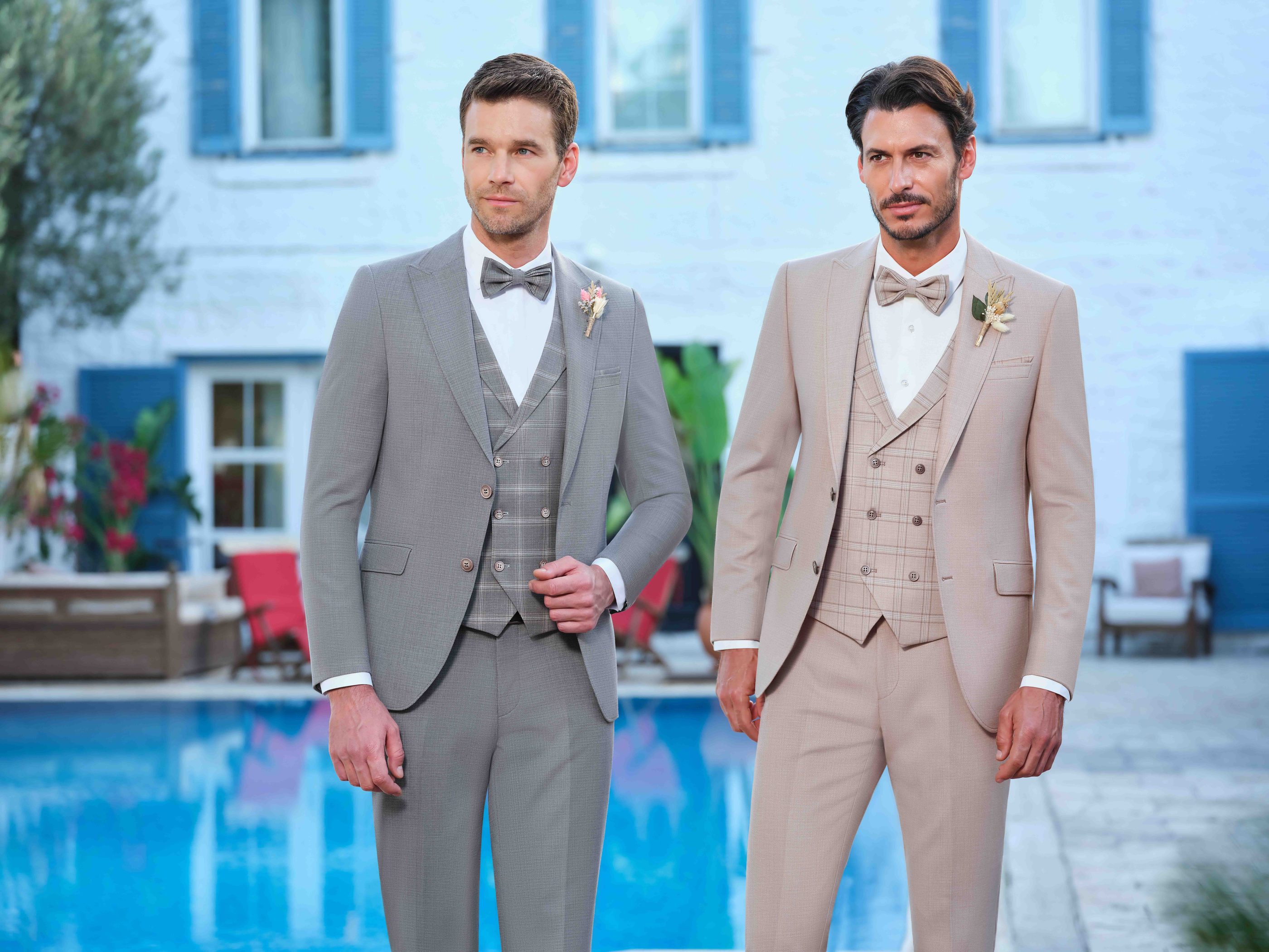 Zwei Männer tragen elegante Hochzeitsanzüge mit Fliegen am Pool vor weißem Haus mit blauen Fensterläden.