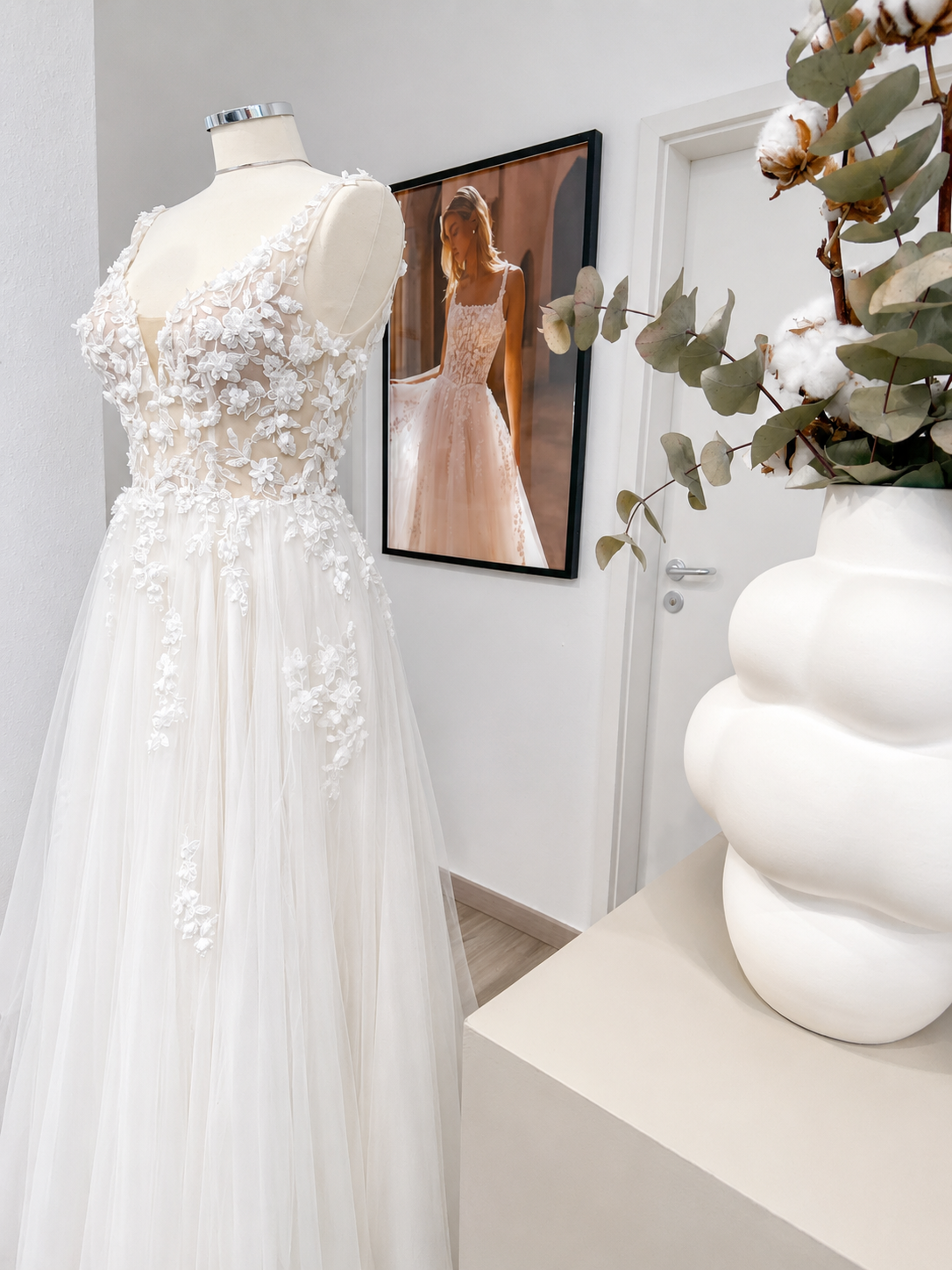 Elegantes weißes Spitzenhochzeitskleid auf Schaufensterpuppe neben Vase mit Baumwollzweigen.